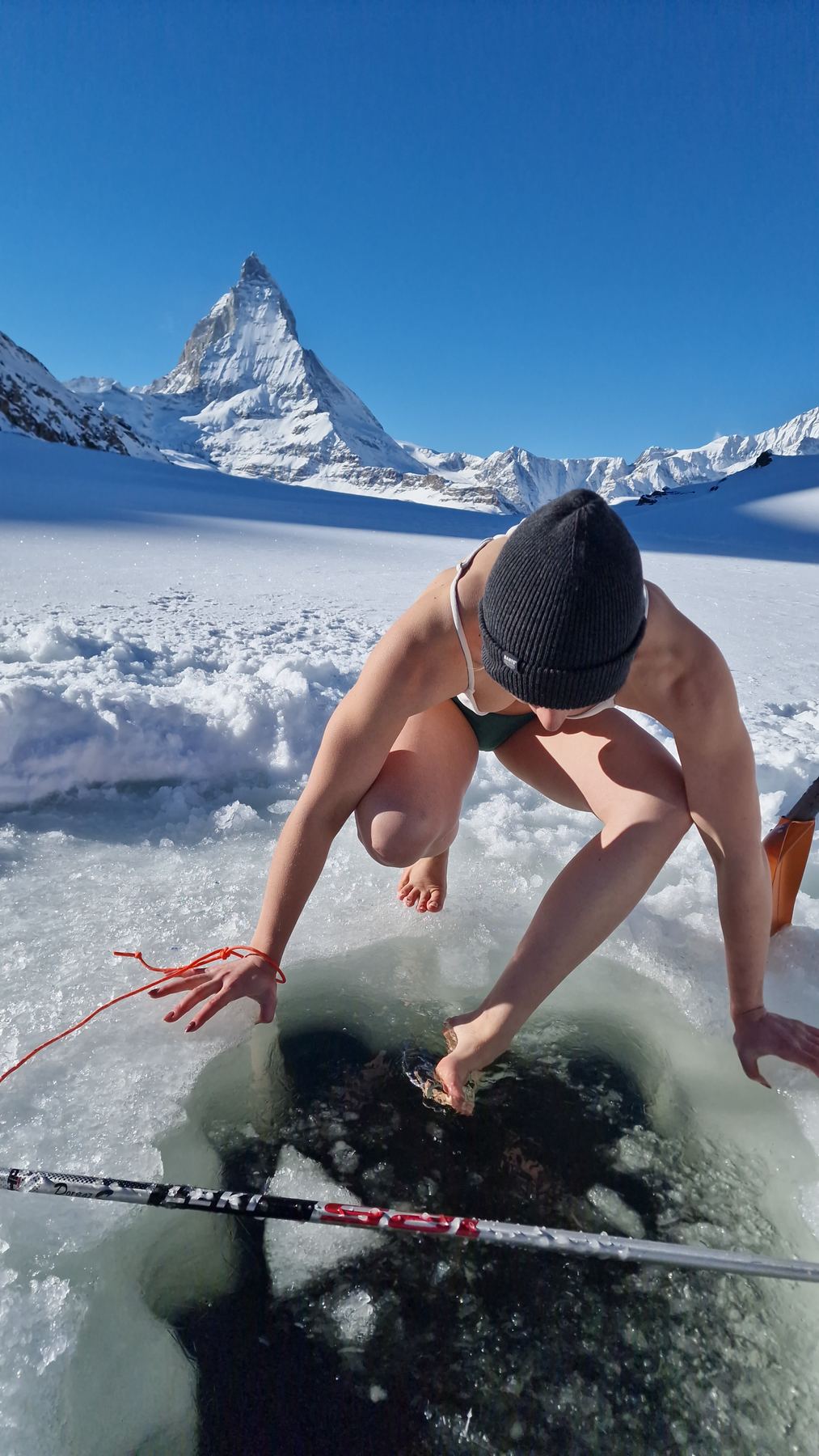A participant lowers herself into a hole in the ice of a frozen lake, the Matterhorn rising in the background.