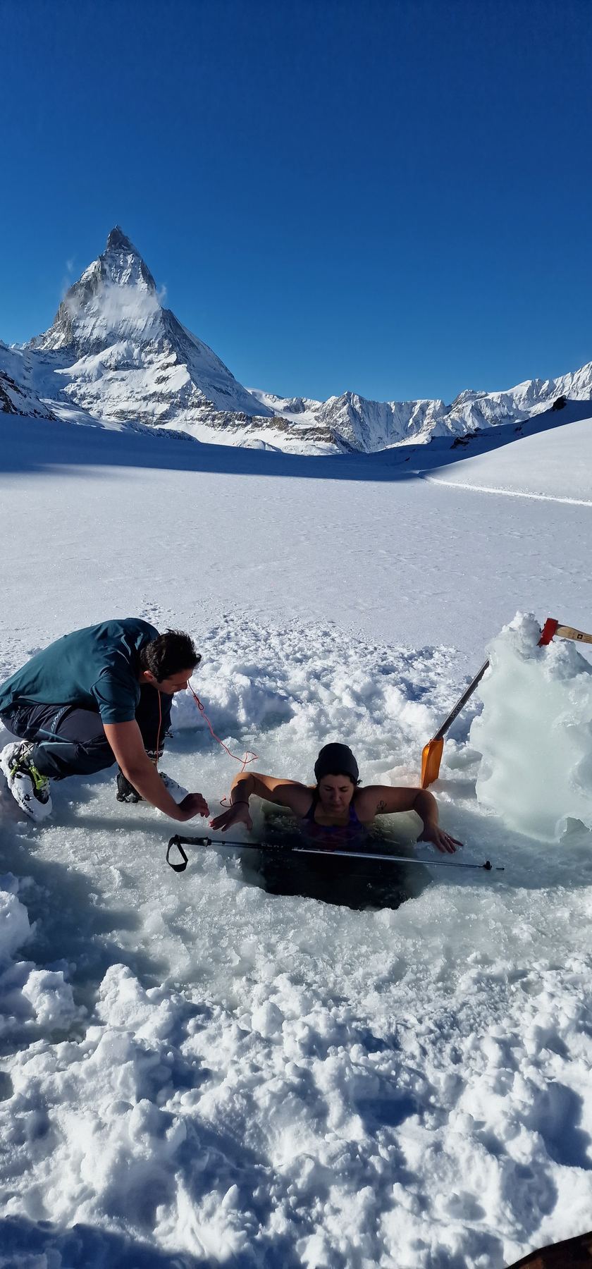A guide crouches at the edge of a hole cut in a frozen lake, encouraging a participant in the water, with the Matterhorn behind them.