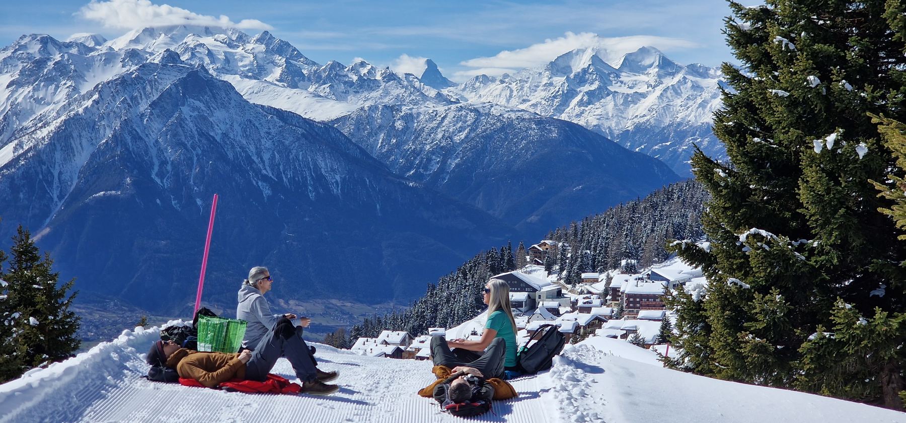 Small group sitting quietly on a groomed snow ridge above a village, facing a panorama of snowy peaks.