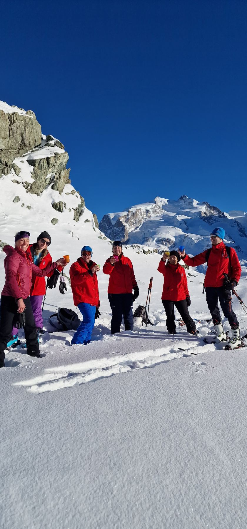 Six smiling ski tourers in red jackets raise colorful cups in a toast on a sunlit snowfield below Monte Rosa.